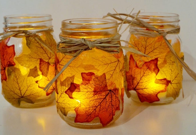 Mason jars decorated with leaves and straw bows each hold a lit candle.