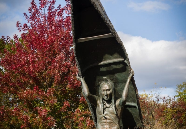 A statue of an Indigenous person portaging a canoe, surrounded by tall grasses.