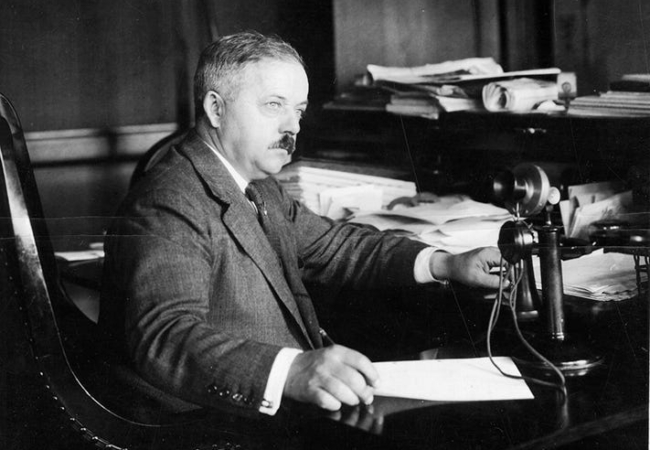 A black and white photo of F.A. Seiberling, a man in a dark suit with a thick mustache, sitting at a cluttered desk with an old fashioned phone in front of him.