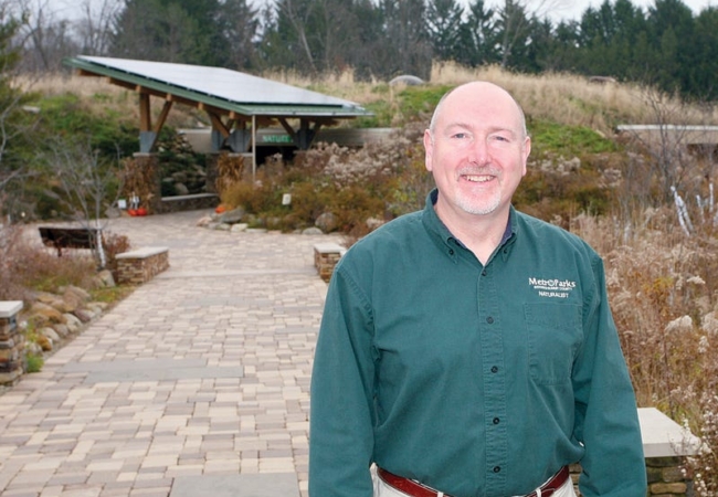 Mike Greene, dressed in a green Metro Parks naturalist shirt, stands on the path in front of Nature Realm Visitors Center, smiling at the camera.