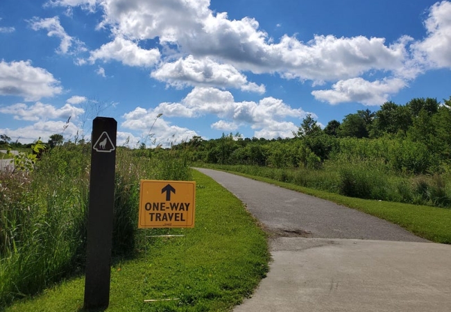 A yellow one-way travel sign stands between a Coyote Run Trail sign and the paved trail.