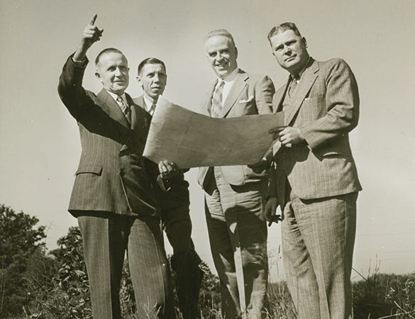 A black and white photo of four men standing in a field, holding a large paper and looking into the distance.