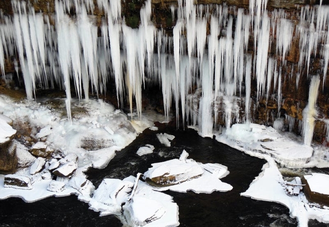 Large icicles hang from rocks along a snowy and partially frozen river below.