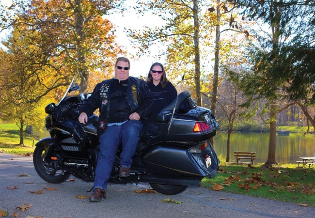 Two people and a motorcycle in front of a pond. One person is sitting on the motorcycle and the other standing behind it.