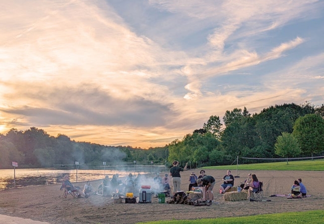A group of visitors sit around a campfire on a sandy beach next to Silver Creek Lake.