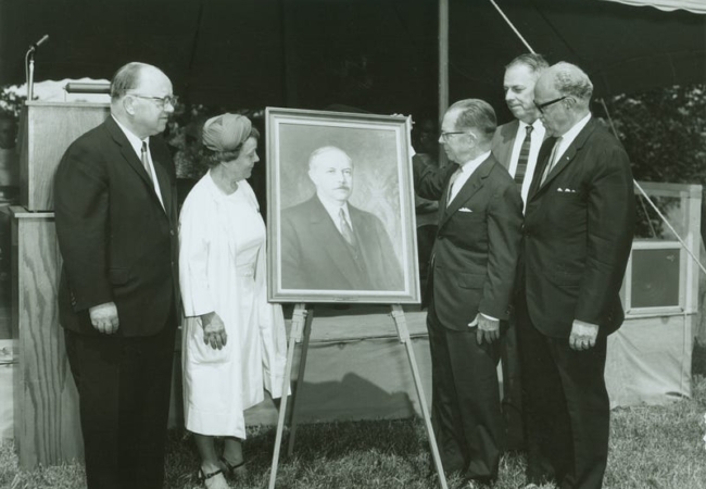 A black and white photo of five people standing around a large photo of F.A. Seiberling.