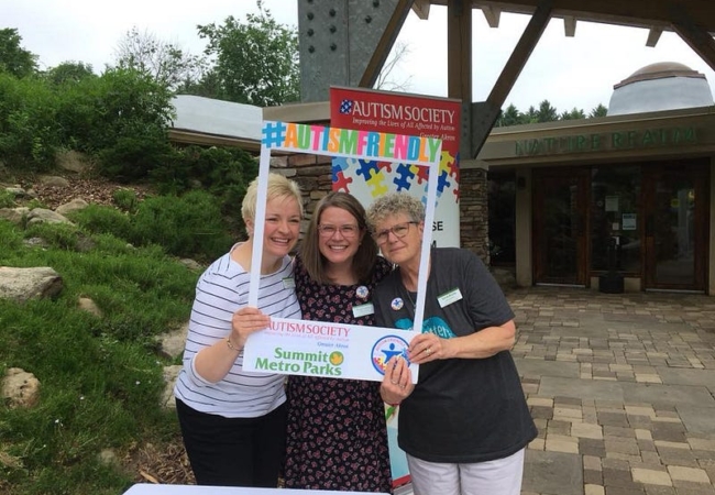 Three women smile at the camera while holding a large photo frame reading #AutismFriendly.