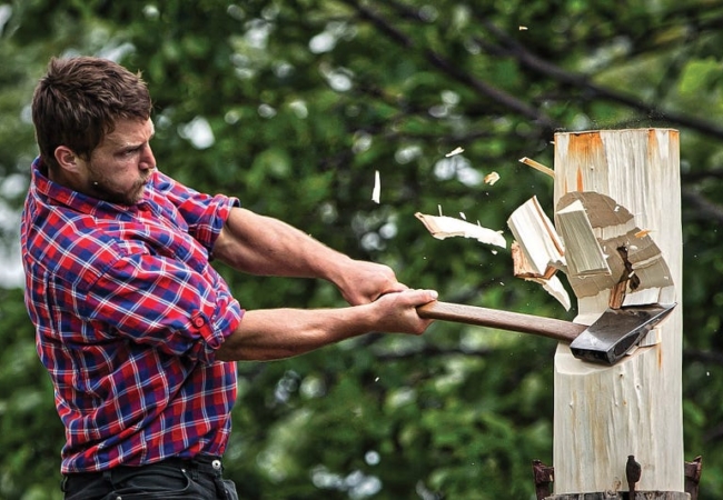 A man swinging a axe, which has made contact with a wooden post. Wood pieces are flying away from the post due to the impact.