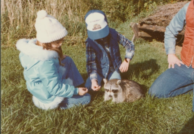 Two kids sit with a raccoon, used many years ago as an ambassador animal.