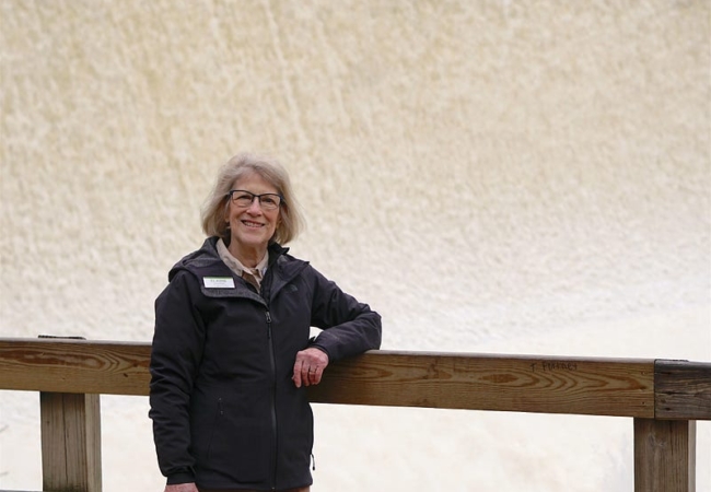 Elaine Marsh leans against a wooden railing. Water rushes over the Gorge Dam in the background.