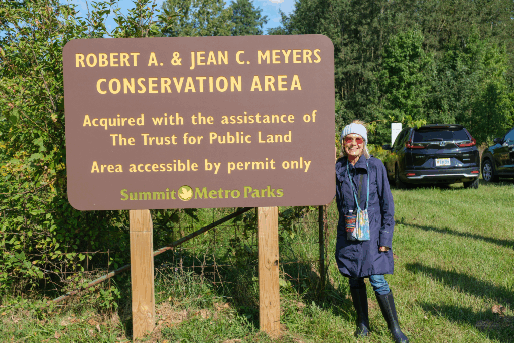 Marilyn Buckey smiles and poses next to a sign for the Robert A. & Jean C. Meyers Conservation Area located at Long Lake.