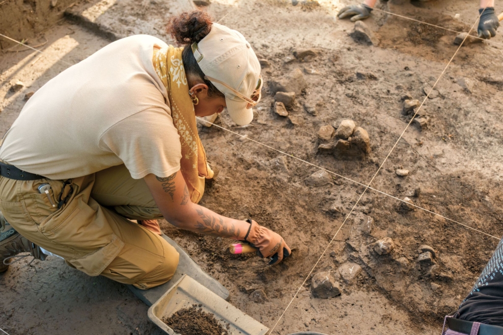Cultural Resources Seasonal Field Technician Zoe Brown kneels next to the 2025 project area, scraping dirt with a tool as she excavates