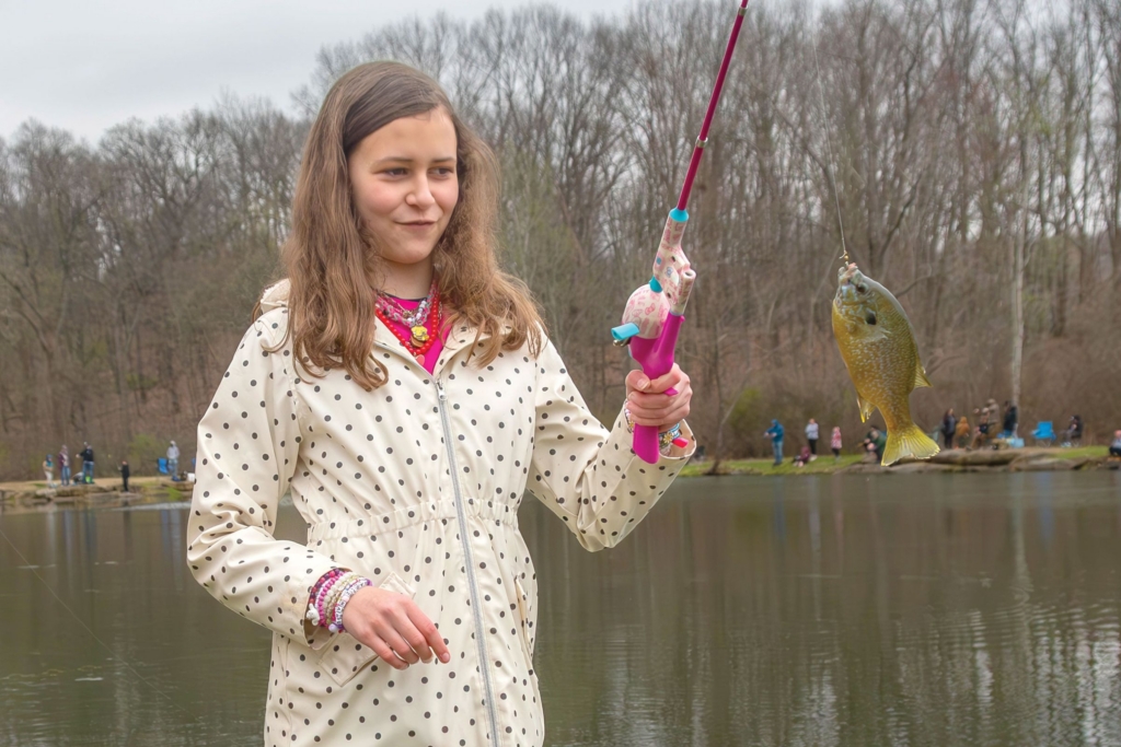 a girl in a polka dotted rain jacket holds a pink fishing pole with a fish hanging from the hook