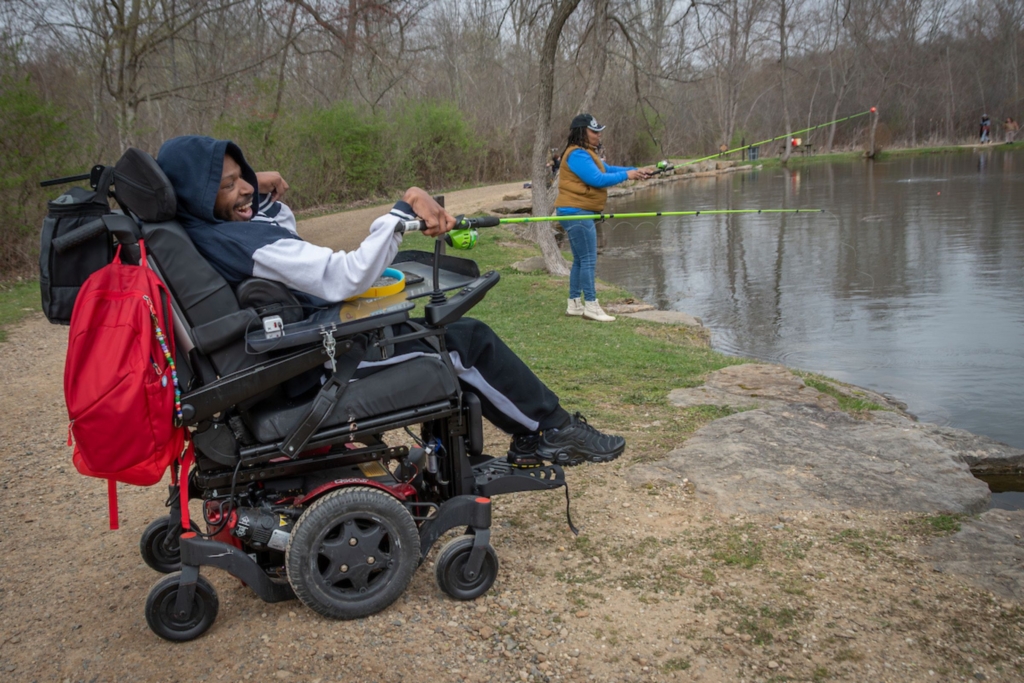 a man in a power wheelchair and a woman standing near him fish at Little Turtle Pond on a spring day