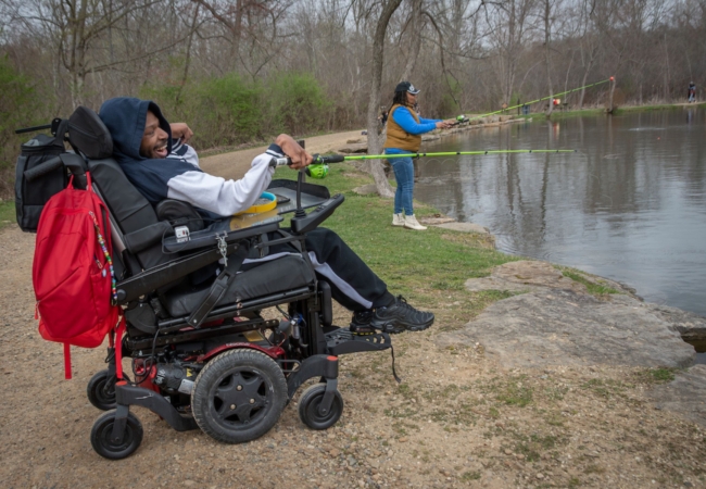 a man in a power wheelchair and a woman standing near him fish at Little Turtle Pond on a spring day