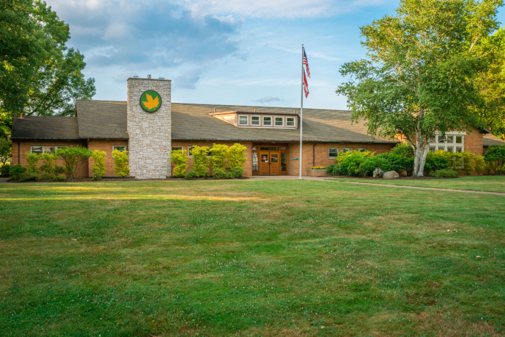 A front-facing view of a lodge sitting on green grass surrounded by green trees and a blue sky