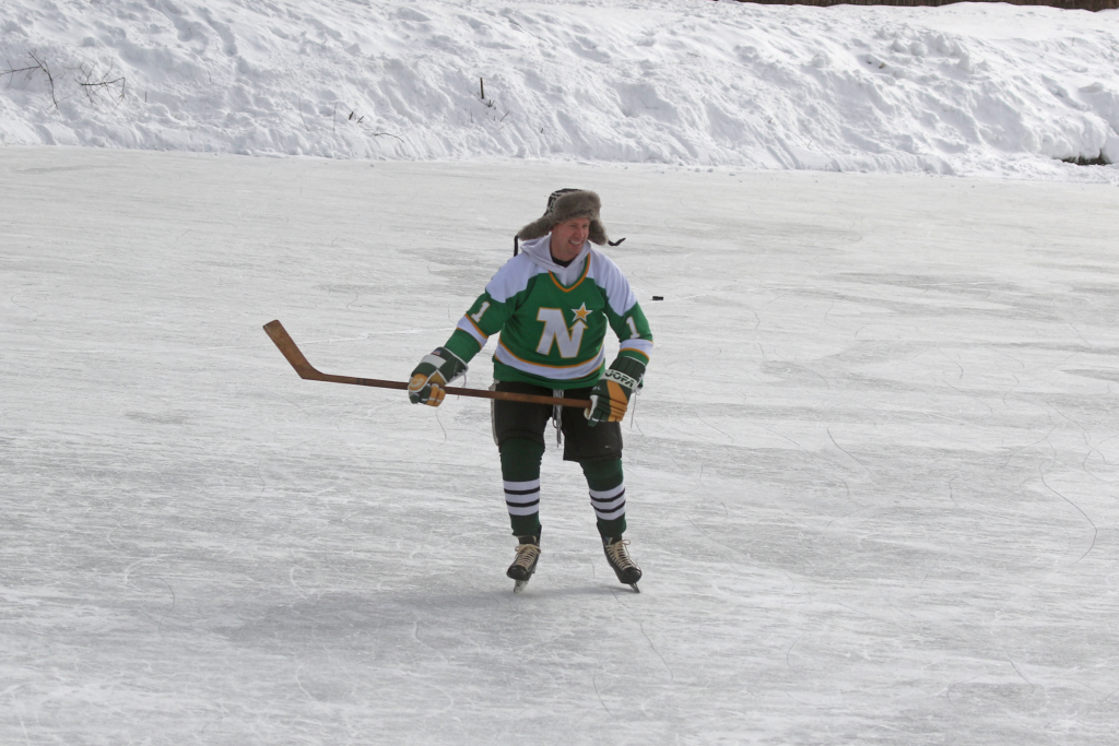 a man in a green hockey jersey, other hockey gear and fur hat skates across Brushwood Lake holding a hockey stick