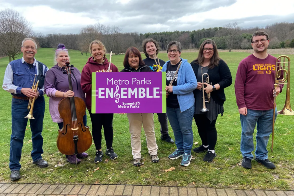 Marketing Assistant Christy Counterman, holding a "Metro Parks Ensemble" sign, with members of the Metro Parks Ensemble during a practice at Goodyear Heights Metro Park.