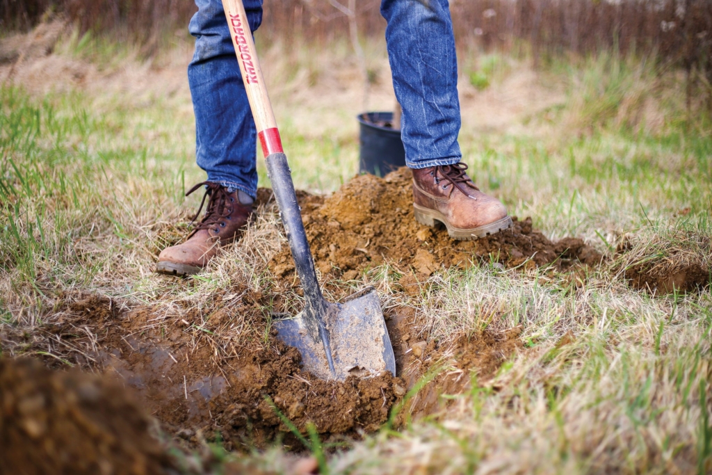mid-shot of person in jeans and boots using a shovel to dig a hole in dirt to plant a tree near Nimisila Creek