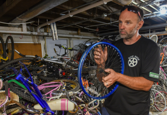 a person wearing a black t-shirt holds a bicycle wheel with bikes and parts in the background