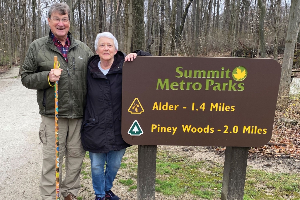 Sam, holding his hiking staff covered in shields, and Anne - both wearing coats - stand next to a trailhead sign at Goodyear Heights Metro Park in late fall