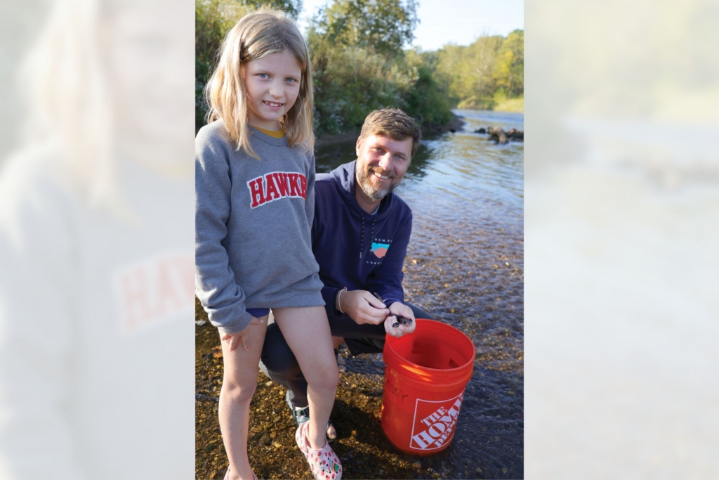 a daughter stands next to her father, who is kneeling behind a 5-gallon bucket and holding a fingerling lake sturgeon