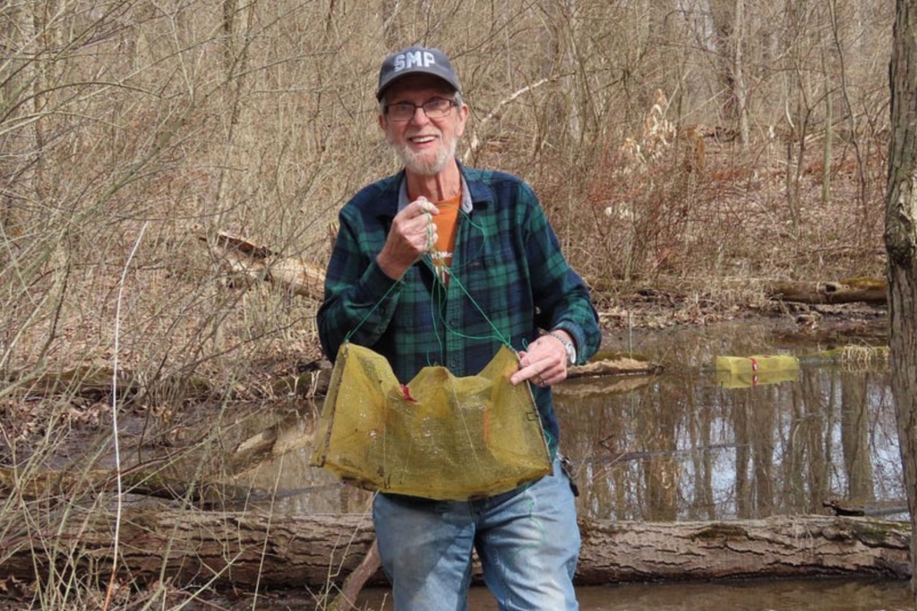 Volunteer Al Casanova stands at a vernal pool, holding a trap used to collect salamanders for surveying. In the background, a trap is set in the vernal pool.