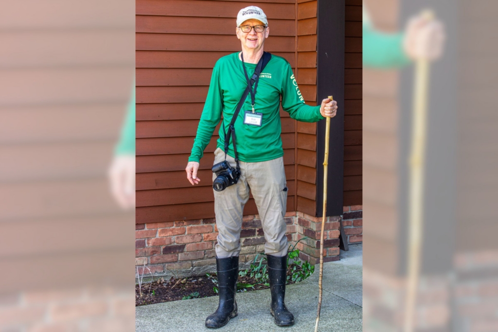 Volunteer Bob stands outside a Metro Parks building, holding a walking stick with a camera slung around his body
