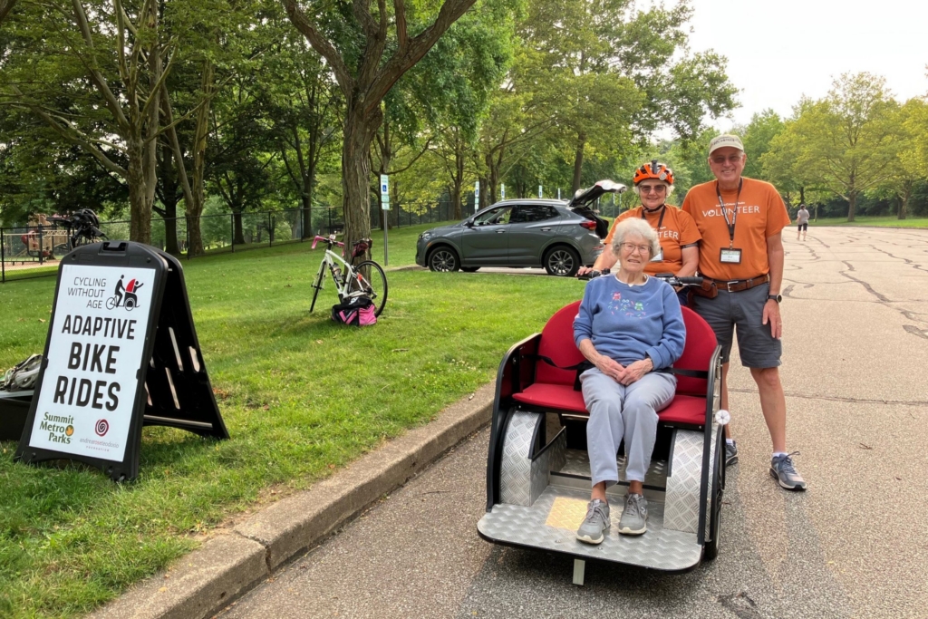 Judy is wearing a helmet while sitting on a trishaw bike with a passenger in the front. Ed stands next to her. Everyone is smiling, ready to enjoy the ride. 