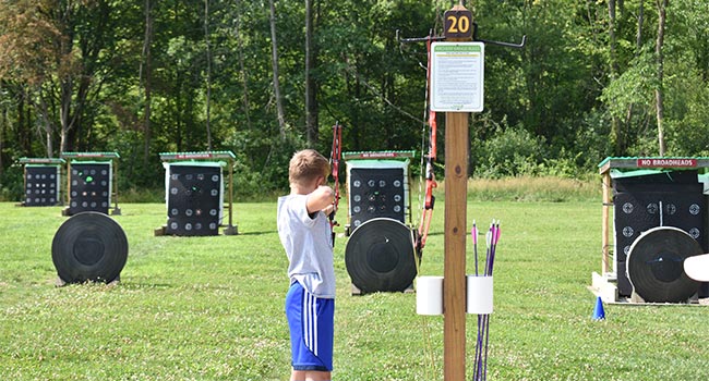 A boy aims a bow and arrow toward a field of archery targets.