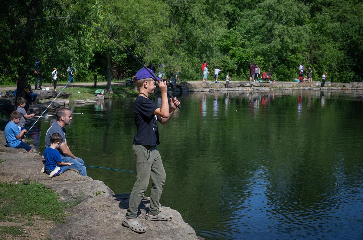 A boy stands at the edge of a fishing pond preparing to cast a reel. Other children fishing line the edge of the pond.