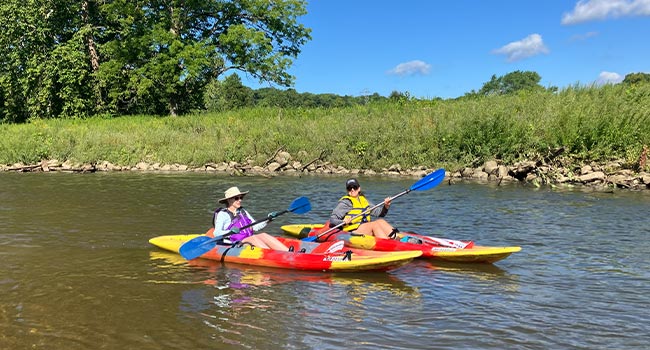 Two kayakers smiling while paddling on a calm river.