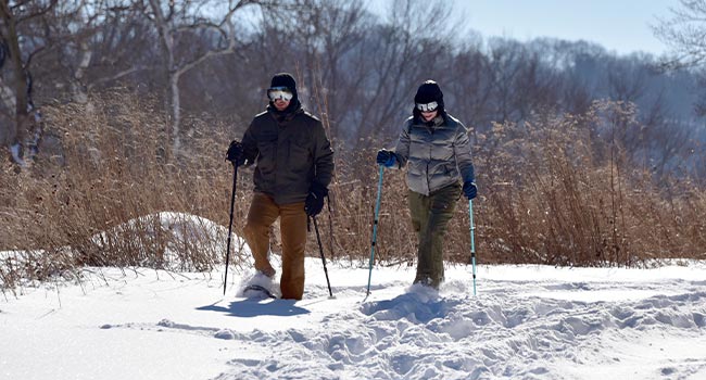 Two people, bundled up in winter gear, snowshoeing along a snow-covered trail.