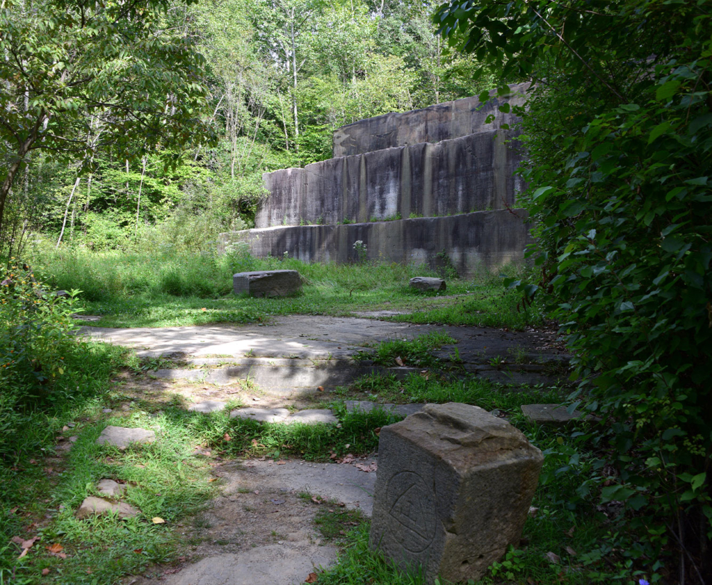 A stone path leads to a layered stone quarry. Both are surrounded by green grass, trees and other foliage.