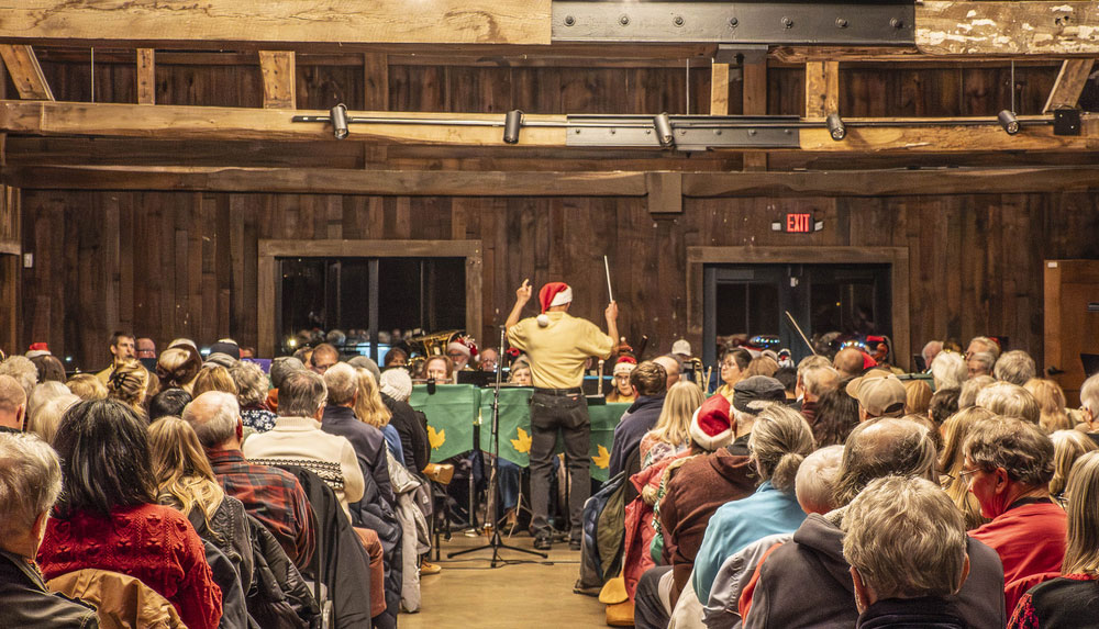 Divided by an aisle, people sit watching a person in a yellow shirt and red and white santa hat conducting a band in a lodge.