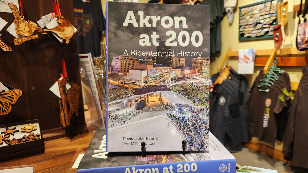 A book sits on a book stand in a gift shop. The title is "Akron at 200: A Bicentennial History."