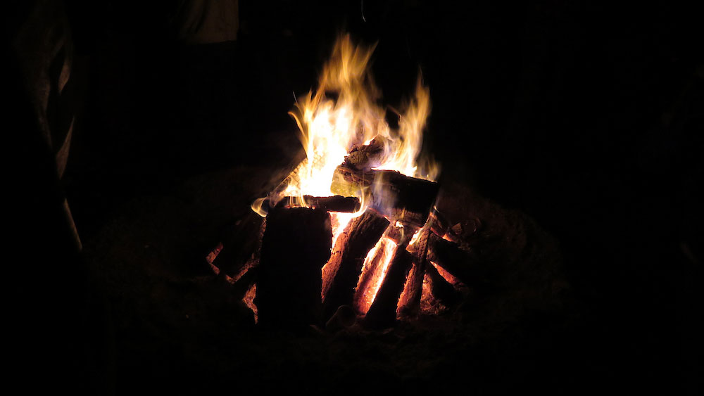 Bright orange and yellow flames rising from a pile of burning logs in a dark setting.