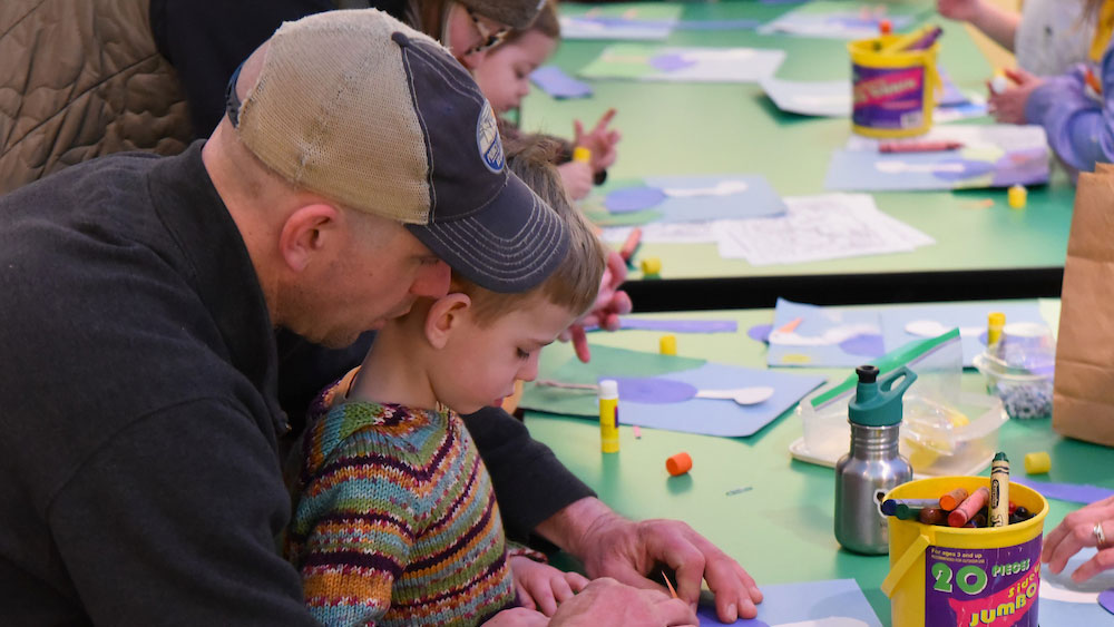 Adult and child working together on a craft project at a green table filled with art supplies and paper cutouts