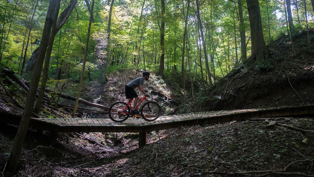 A mountain biker rides across a wooden bridge on a forest trail, surrounded by tall trees and dappled sunlight