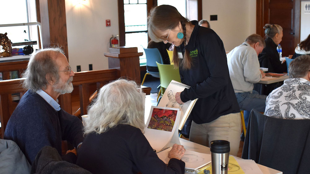 A person standing and showing artwork in a sketchbook to two seated people indoors