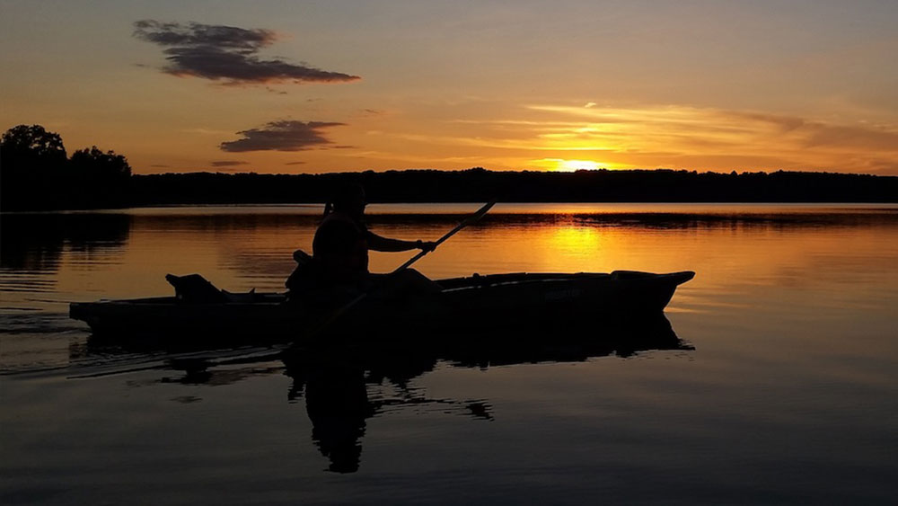 A kayaker paddling across a body of water while the sun sets in the distance.