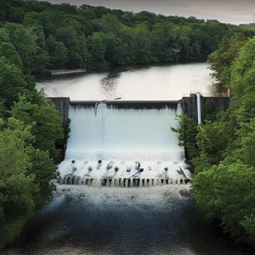 White water rushes from a wide dam pool, over a large concrete dam, and to the river below. The river and dam are tightly surrounded by green trees.