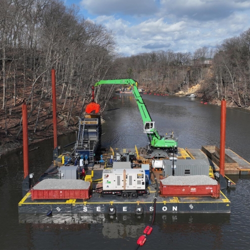 A large green crane and other dredging equipment on a black platform barge float at the surface of a river of dark water. Bare trees line the riverbanks and the blue sky is full of white clouds.