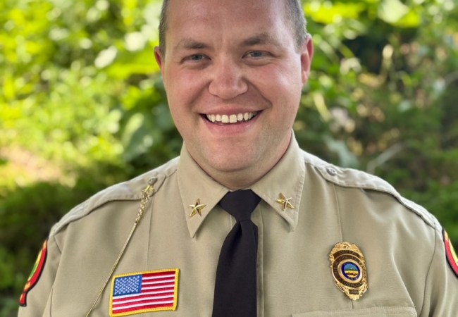 a man dressed in a khaki colored uniform shirt and black tie smiles with greenery and a blue sky in the background