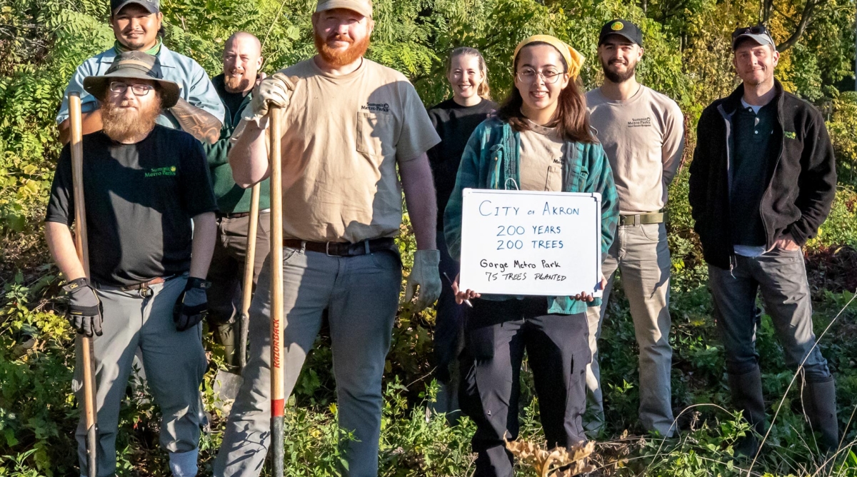 A group of Summit Metro Parks staff pose wearing working gear. One is holding a sign that says, "City of Akron, 200 years, 200 trees. Gorge Metro Park, 75 trees planted."