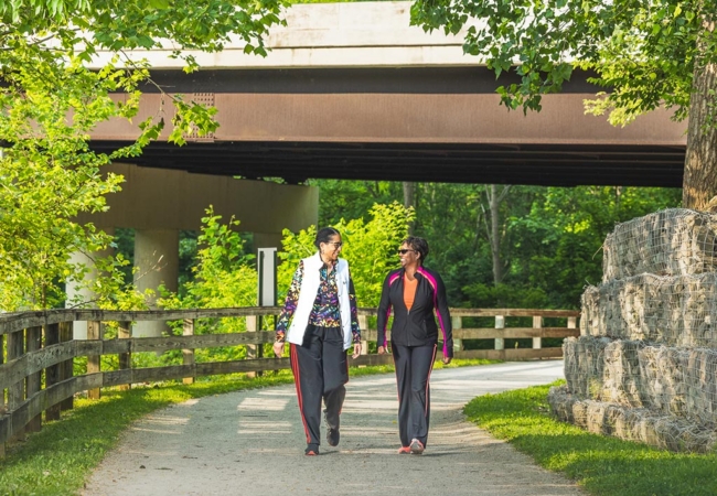 Two women walk on a flat trail surrounded by rocks and trees. Smiles are on both of their faces.