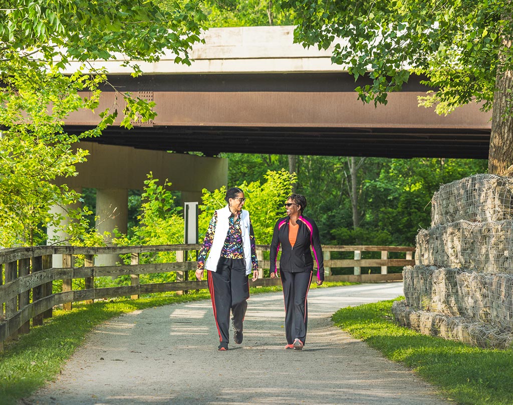 Two women walk on a flat trail surrounded by rocks and trees. Smiles are on both of their faces.