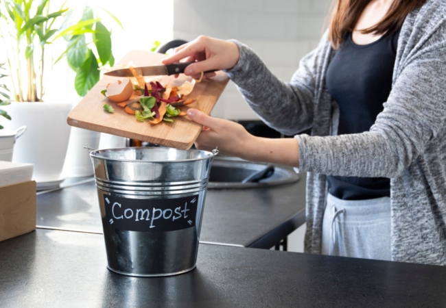A white woman scrapes food scraps from her cutting board into a tin bucket labeled "Compost" in her kitchen