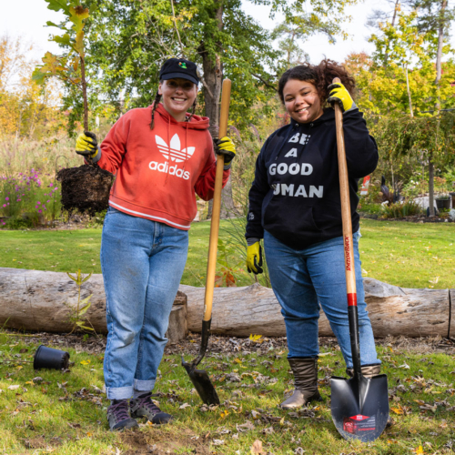 Two woman wearing work gloves and holding shovels. One woman is also holding a plant in a nursery pot.