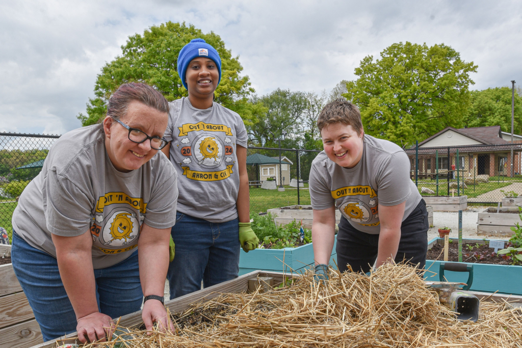 Three people in matching gray Out N' About t-shirts lean over a community garden bed and smile at the camera